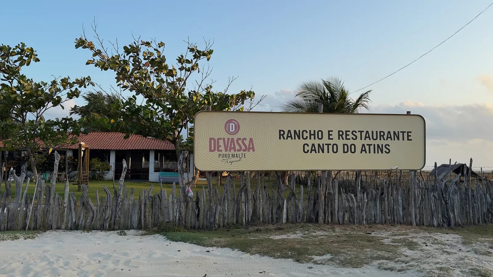 Enseigne sur la clôture en bois avec le nom Rancho e Restaurante Canto do Atins, avec le sable de la plage devant